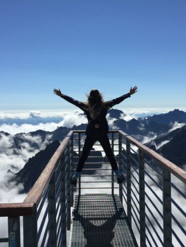 Person Standing on Hand Rails With Arms Wide Open Facing the Mountains and Clouds