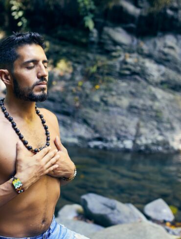 topless man wearing black beaded necklace and blue denim shorts standing on rocky shore during daytime