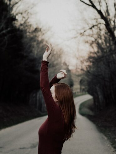 selective focus photography of female raising hands in the middle of the road surrounded by withered trees