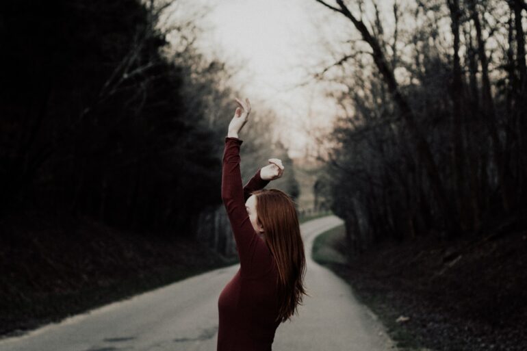 selective focus photography of female raising hands in the middle of the road surrounded by withered trees