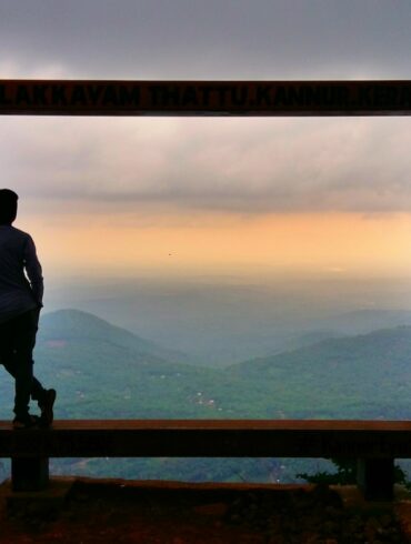 man in black jacket standing on brown wooden fence looking at mountains during daytime