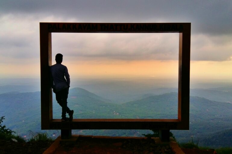 man in black jacket standing on brown wooden fence looking at mountains during daytime