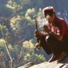 man sitting on rock formation near cliff while holding smoking pipe during daytime