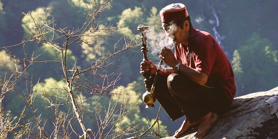 man sitting on rock formation near cliff while holding smoking pipe during daytime