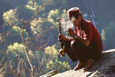 man sitting on rock formation near cliff while holding smoking pipe during daytime