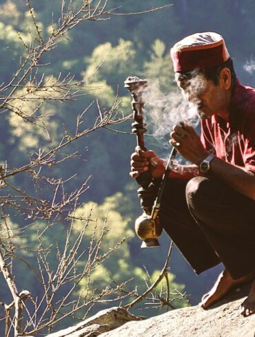 man sitting on rock formation near cliff while holding smoking pipe during daytime