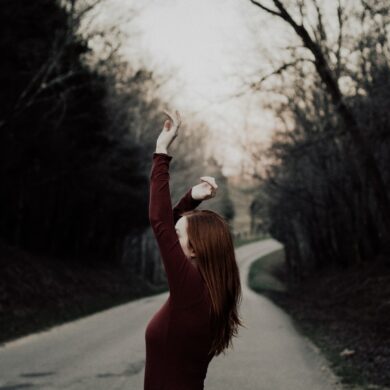 selective focus photography of female raising hands in the middle of the road surrounded by withered trees